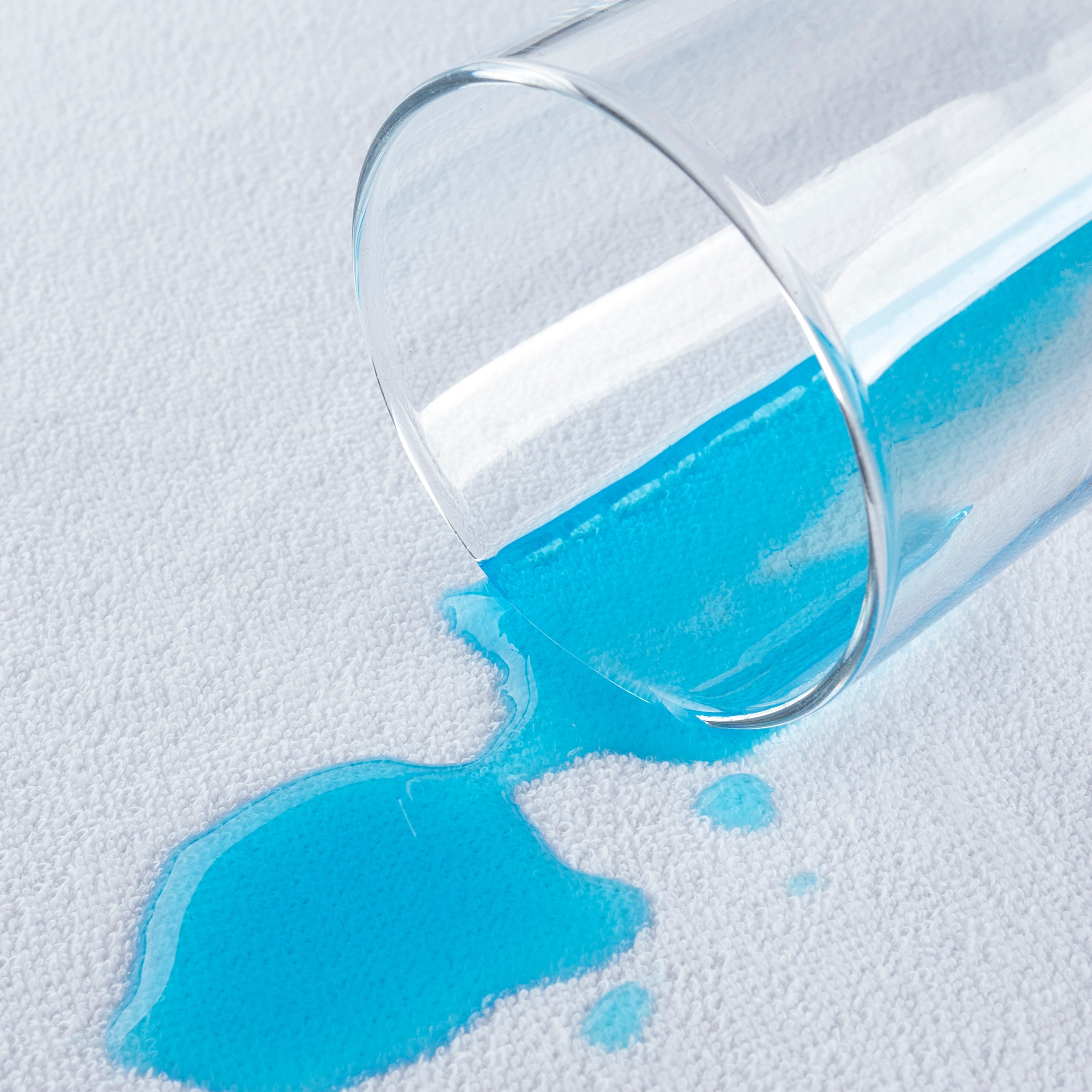 A clear glass tumbler tipped over on a white textured surface, spilling bright blue liquid in a small pool.